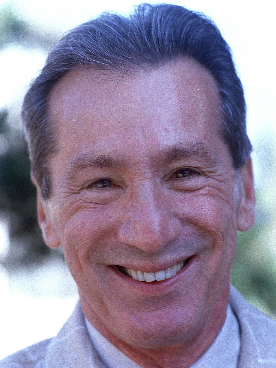 A smiling middle-aged man with dark hair streaked with gray, wearing a light-colored collared shirt, outdoors with blurred greenery in the background.