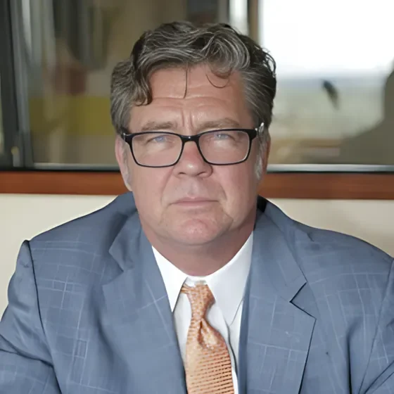 A middle-aged man with gray hair and glasses, wearing a blue suit and a patterned tie, sitting indoors in front of a window.