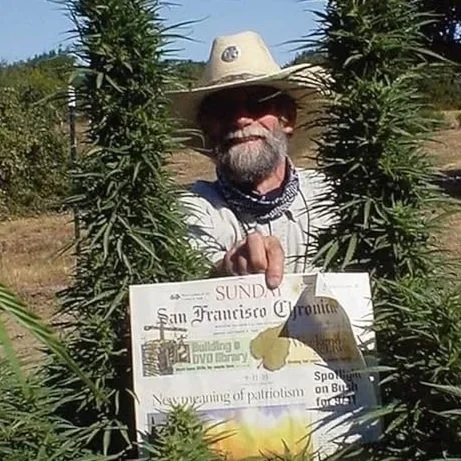 A man with a beard wearing a cowboy hat, sunglasses, and a bandana around his neck, holding a sign inside a marijuana plant.