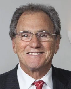A smiling middle-aged man with gray hair, glasses, wearing a dark suit, white shirt, and red tie.