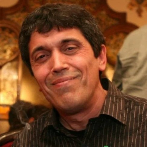 A man with dark hair, wearing a black button-up shirt, smiling in a warmly decorated setting.