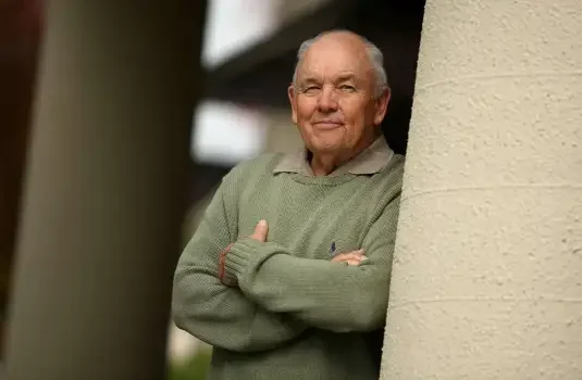 An elderly man with a slight smile, arms crossed, standing outdoors near a beige wall.