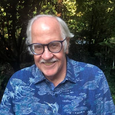 A smiling elderly man with white hair, glasses, and a mustache wearing a blue Hawaiian shirt, outdoors with trees in the background.