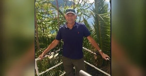 Man standing on a balcony surrounded by green tropical plants, wearing a navy polo shirt, khaki pants, and a cap.