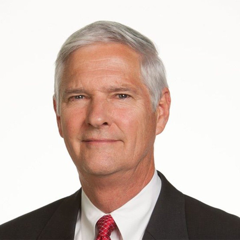 A portrait of a middle-aged man with gray hair, wearing a dark suit, white shirt, and red tie, against a plain white background.