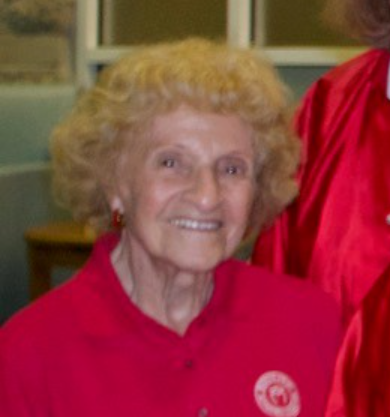 An elderly woman with curly blonde hair smiling, wearing a red shirt, in an indoor setting.