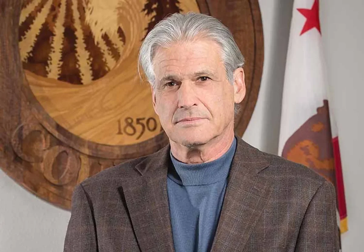 An older man with gray hair wearing a brown blazer and blue turtleneck standing in front of a wooden emblem with '1850' inscribed, and a California state flag in the background.