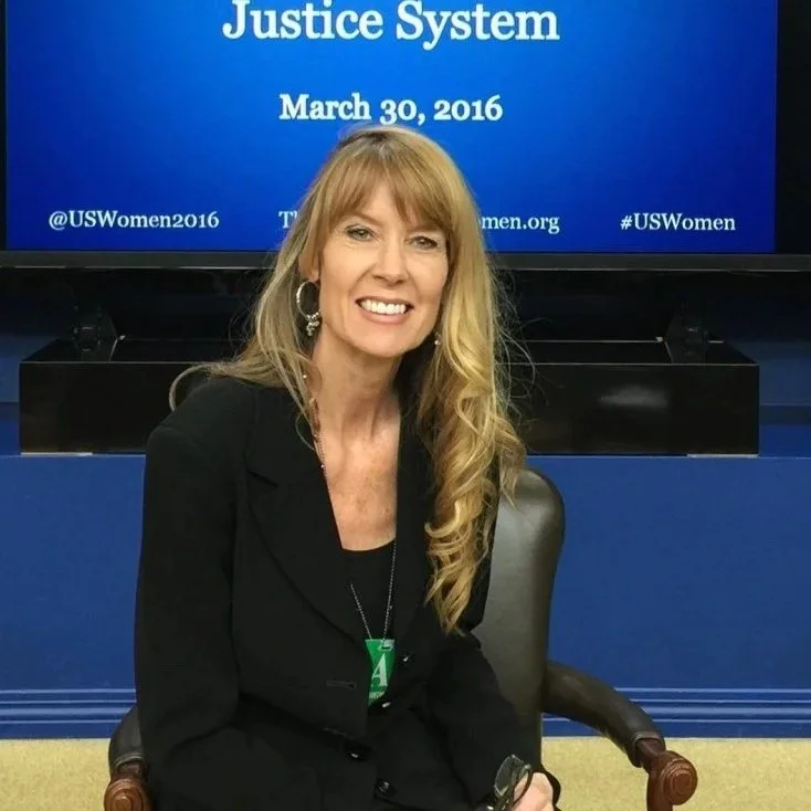 A woman with long blonde hair wearing a black blazer, sitting in a chair, smiling at the camera. Behind her, a blue sign reads, 'Justice System March 30, 2016' with Twitter handles and hashtags related to US Women.