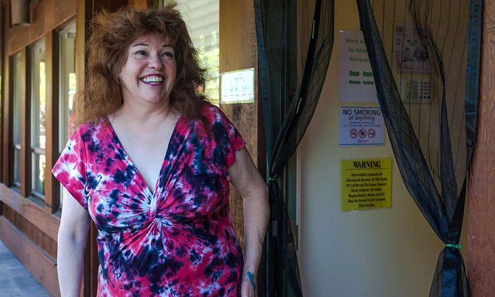 A woman with curly red hair wearing a red, pink, and blue tie-dye shirt smiling outdoors near a wooden building.