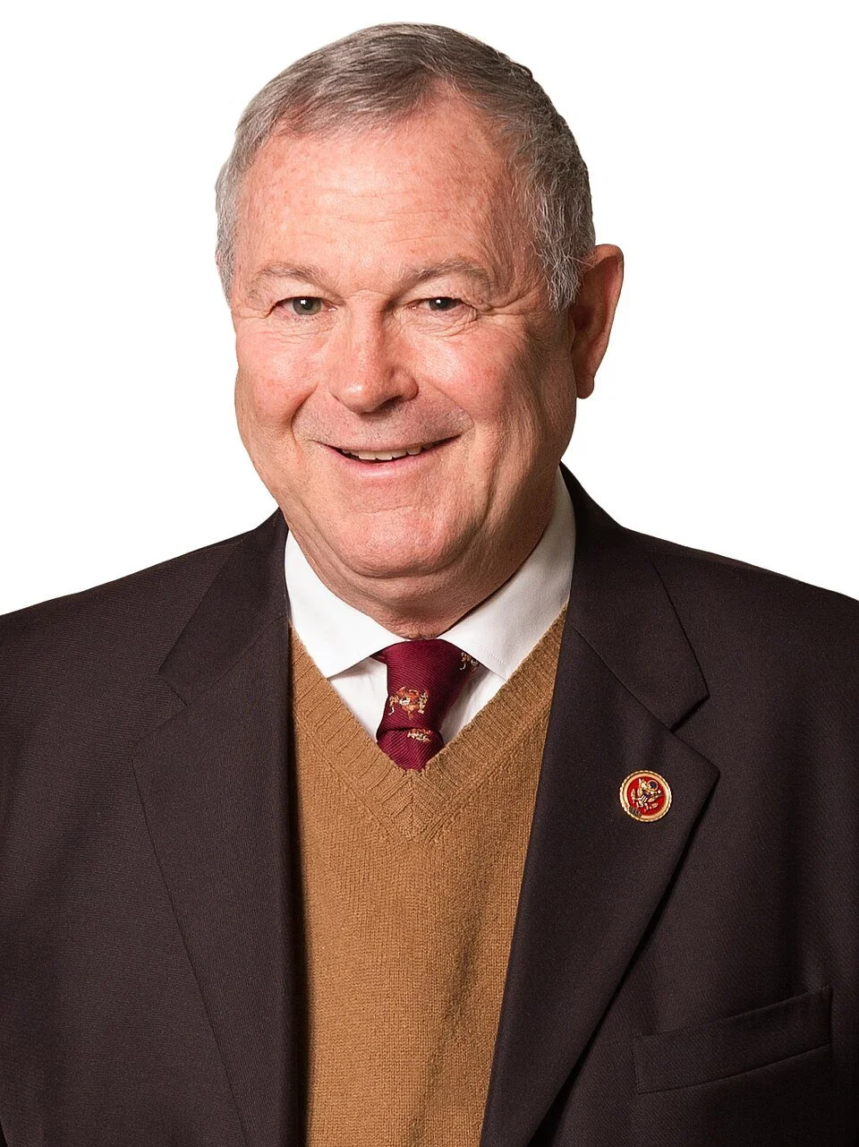 A smiling elderly man wearing a suit jacket, a khaki sweater, a white shirt, and a maroon tie with a small emblem. He has short gray hair and a pin on his lapel.