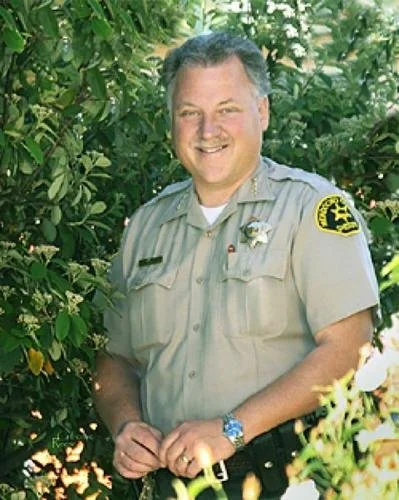A smiling man in a sheriff uniform standing outdoors among green foliage, with patches on his shirt and a badge on his chest.