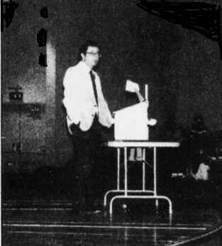 Black and white photo of a man standing next to a table with a box and a small model airplane on it, in a room with a dark background.