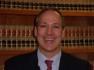 A man in a suit smiling in front of a bookshelf filled with legal books.