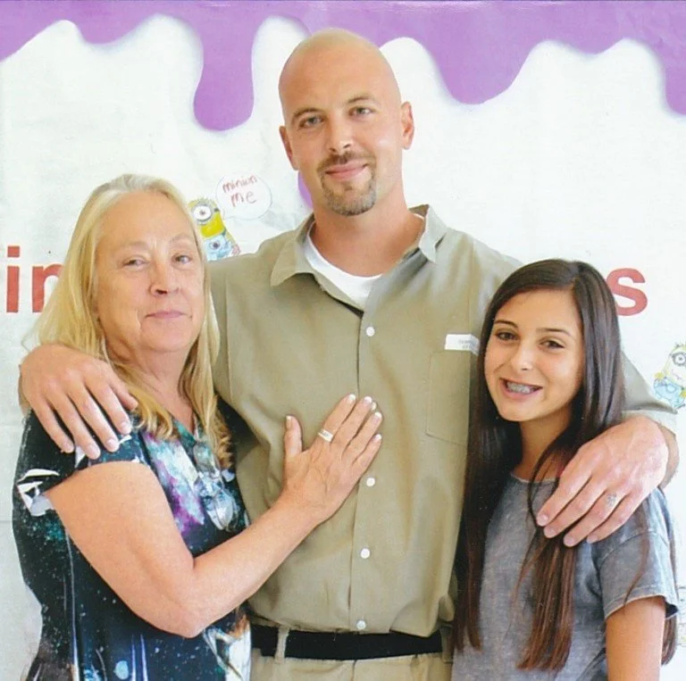 Three people, a woman with long blonde hair, a man with a beard, and a young girl with long dark hair, stand close together and smile, with their hands on each other's shoulders or chest. They are indoors with a purple and white background featuring minion characters and text.