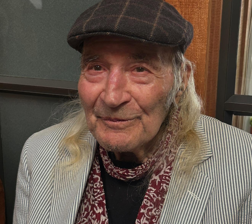 An elderly man with long gray hair, wearing a checkered flat cap, a white striped blazer, and a patterned scarf around his neck, smiling.