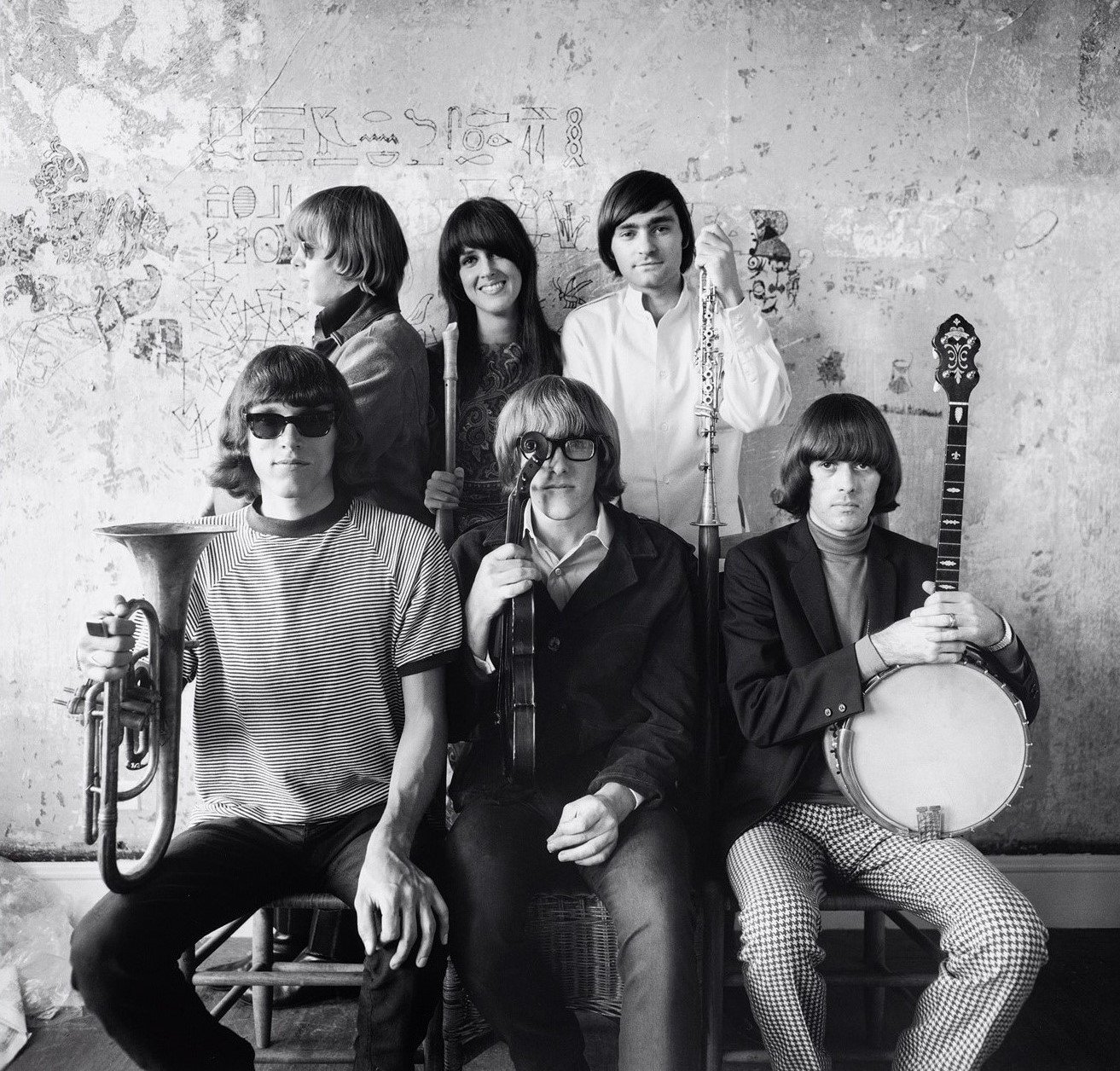 Black and white photo of seven young adults with musical instruments, standing and sitting against a graffiti-covered wall.