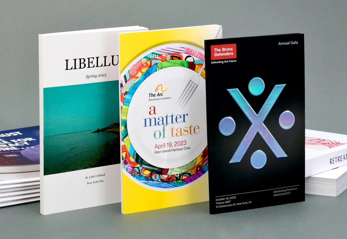Three printed brochures stand upright on a surface. The first is labeled 'LIBELLUS' from St. Luke’s School, NYC, with a blue ocean image. The second is a colorful flyer for 'a matter of taste' event on April 19, 2023, at Glen Island Harbour Club. The third is a black brochure for 'The Bronx Defenders Annual Gala' on October 19, 2023, featuring geometric designs.