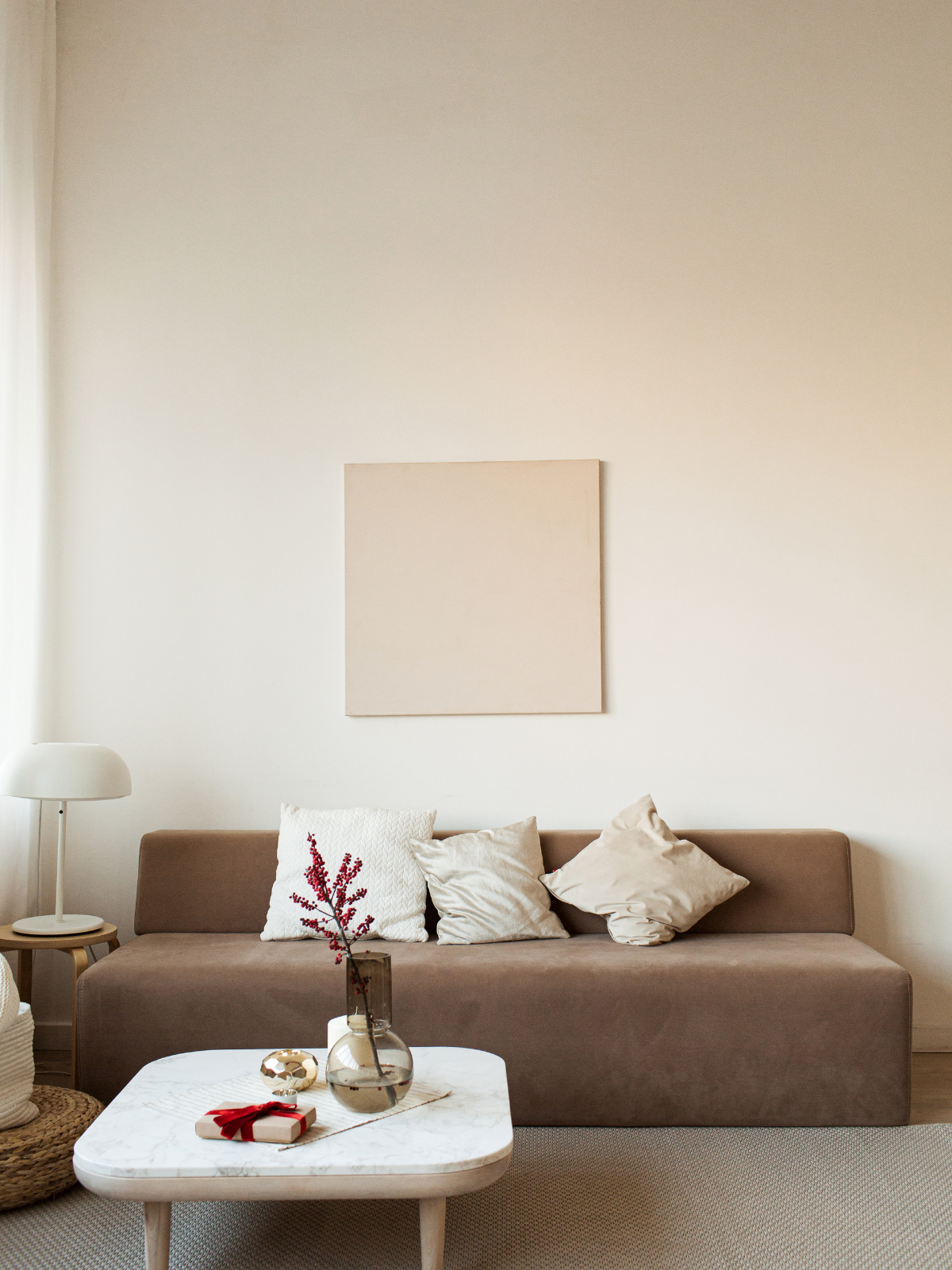 Living room with beige sofa, three pillows, a side table with a lamp, a framed art piece on the wall, beige curtains, and a marble coffee table with a vase, a gift box, and a decorative object.