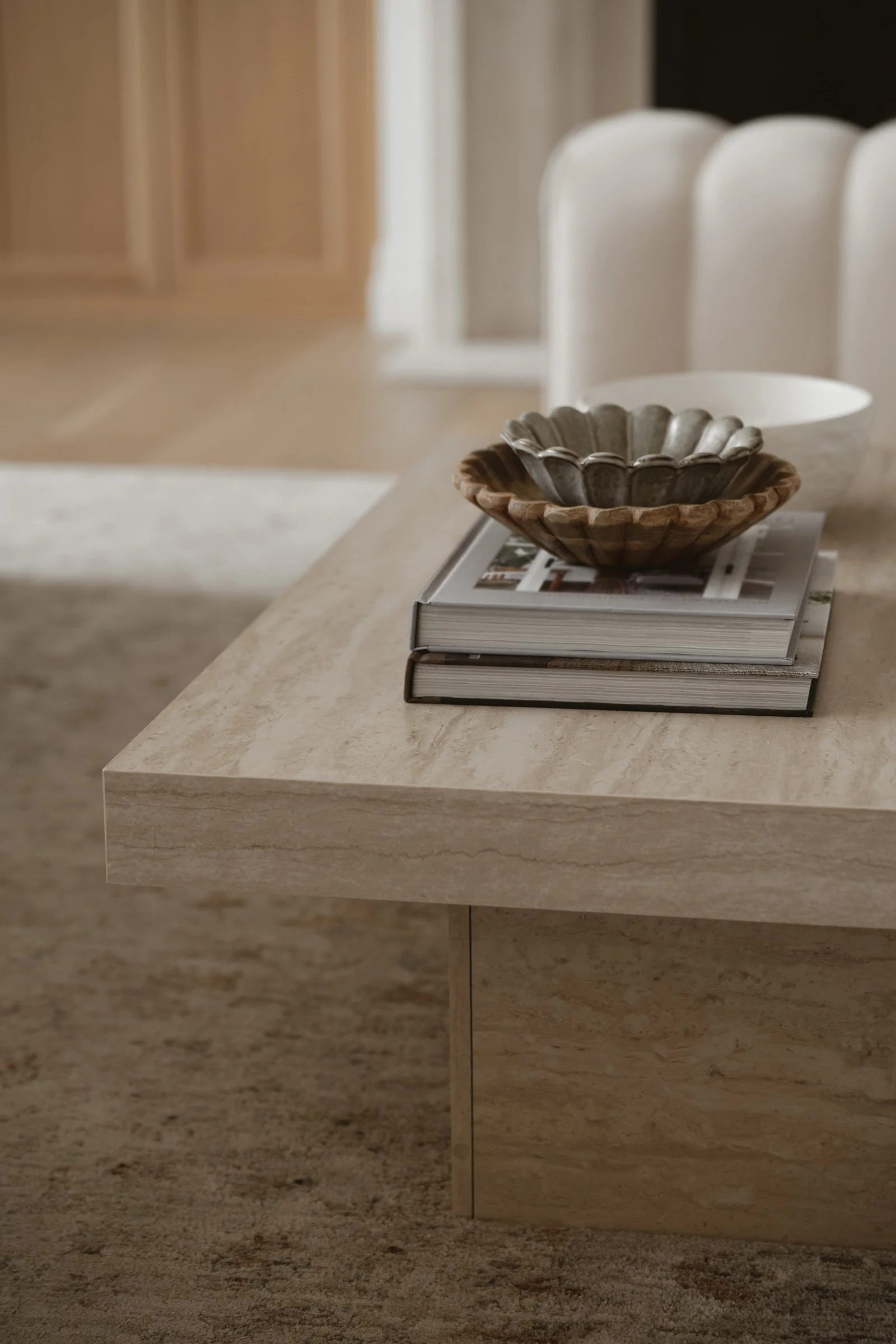 A beige marble table with a wood and metal bowl, a white ceramic bowl, and a closed magazine on top of it.