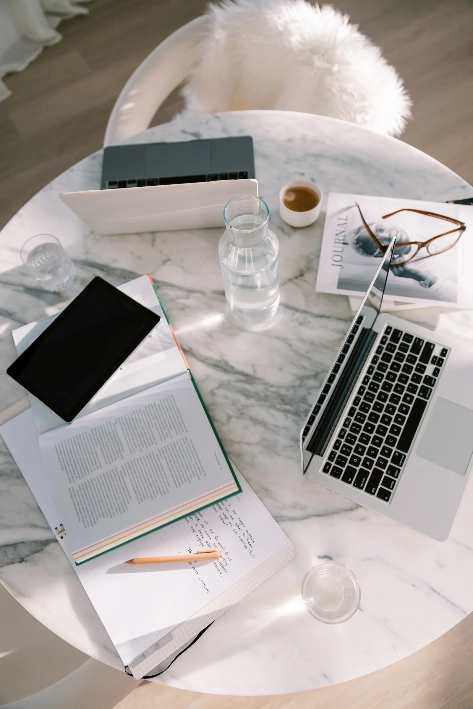 A marble table with an open book, a tablet, two laptops, a smartphone, a notebook with handwritten notes, a pen, a glass of water, a cup of coffee, a magazine, and a pair of eyeglasses.