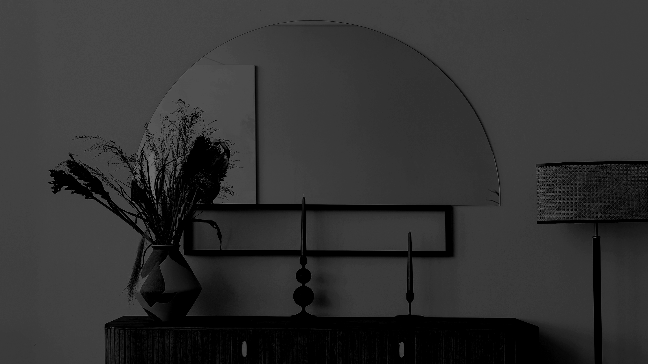 A dark-toned interior with a decorative vase of dried flowers on a sideboard, a rectangular mirror, a half-circle mirror, a candlestick, and a table lamp with a patterned shade.