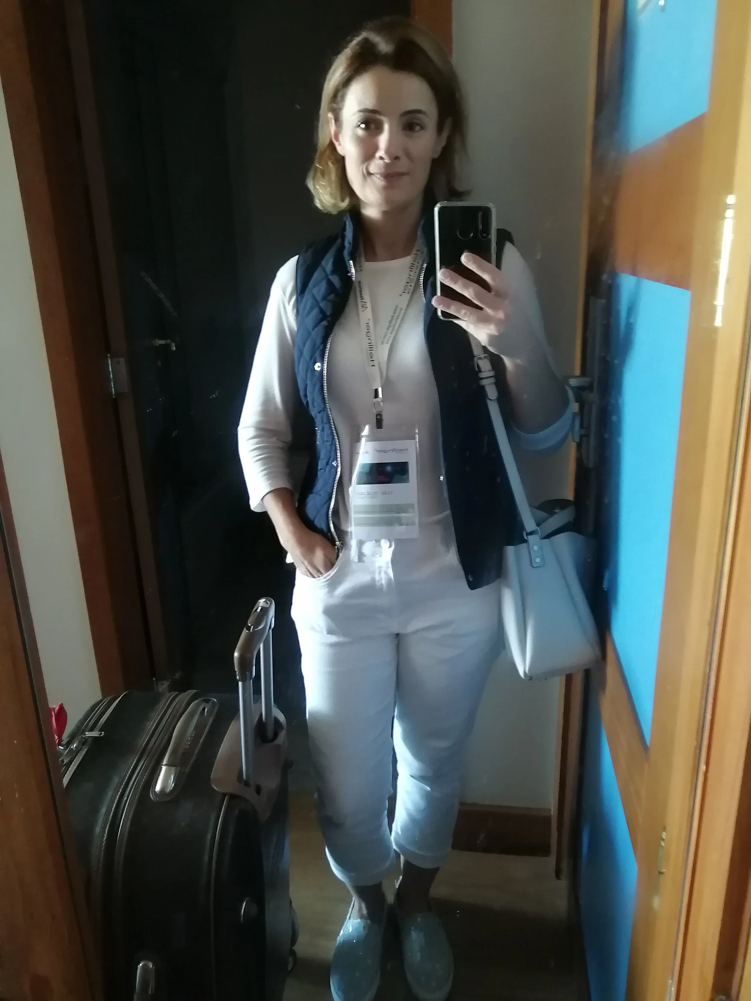 Mulher fazendo selfie em espelho na entrada do hotel, com mala de rodinhas ao lado, vestindo roupa branca, colete azul, tênis cinza e com crachá.