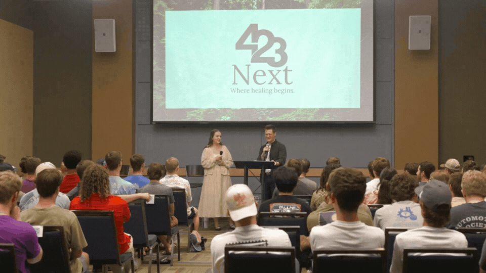 A speaker and a woman with a microphone on stage in front of an audience in a hall with a large screen behind them displaying the logo '423 Next' and the motto 'Where healing begins.'