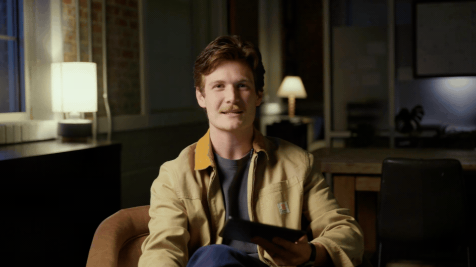 A young man sitting in a cozy, dimly-lit room during evening, holding a tablet.