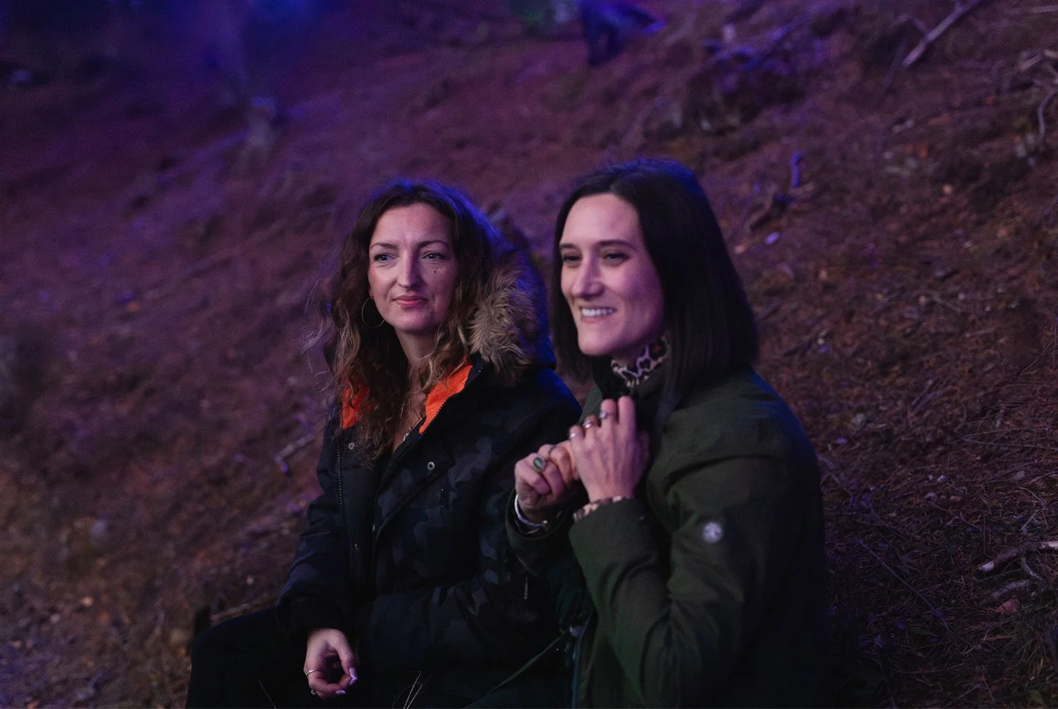 Two women sitting on the ground outdoors at night with cool, blue lighting and trees in the background.