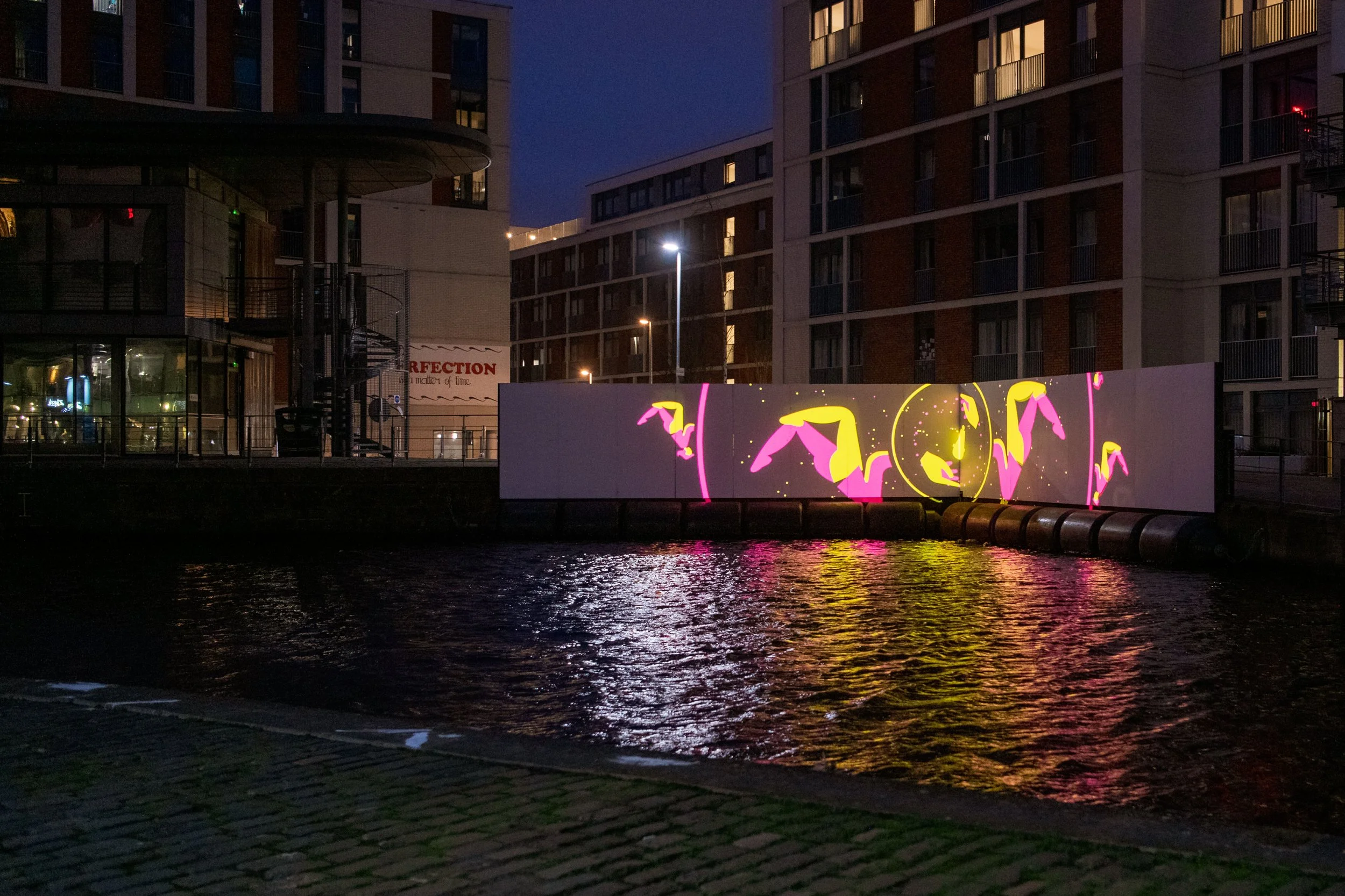 Nighttime view of a city waterfront with a digital art projection on a white wall, showing pink flamingos and yellow circles reflected on the water, surrounded by modern apartment buildings.