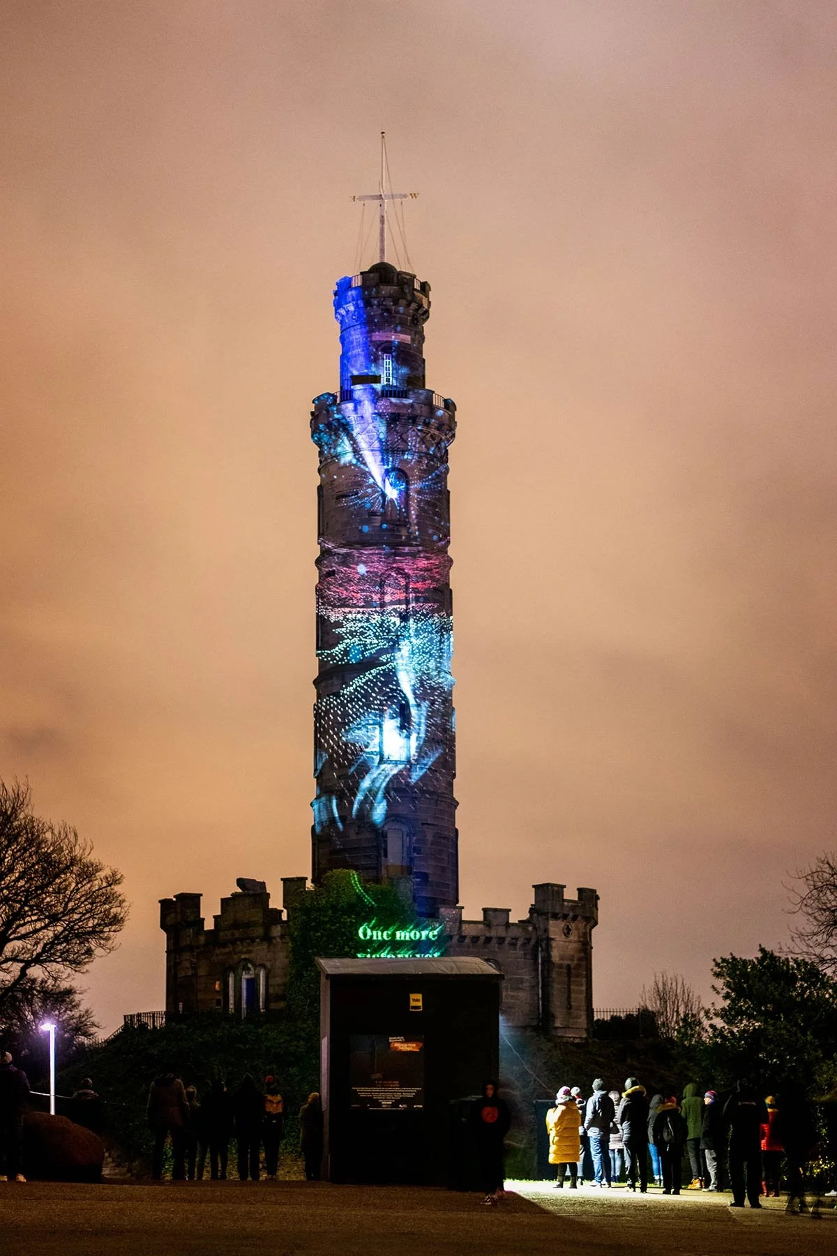 A tall, illuminated tower at night with projections of colorful lights, patterns, and images. People stand at the base, looking up, with a dark sky overhead.