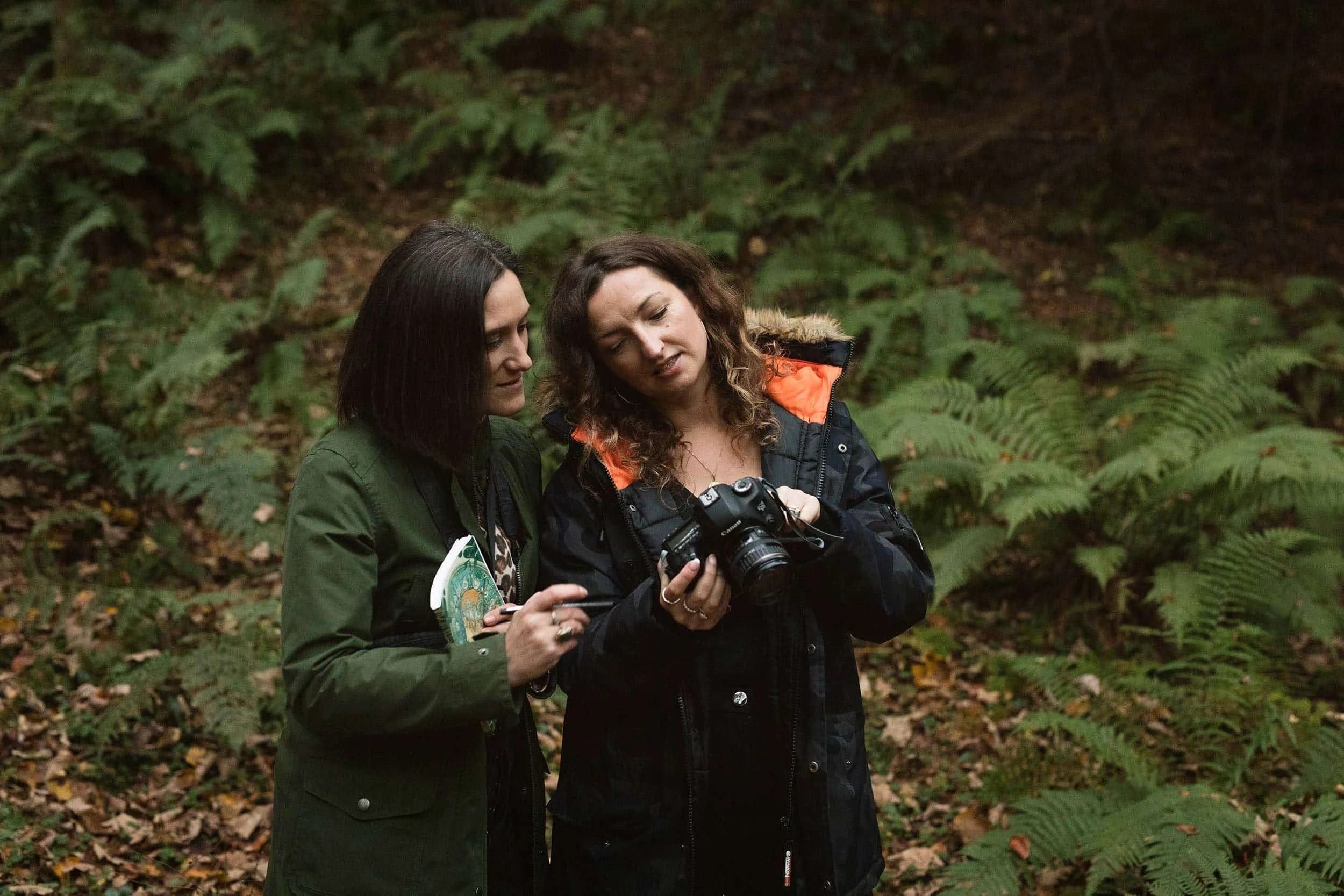Two women standing in a forest, looking at a camera together.