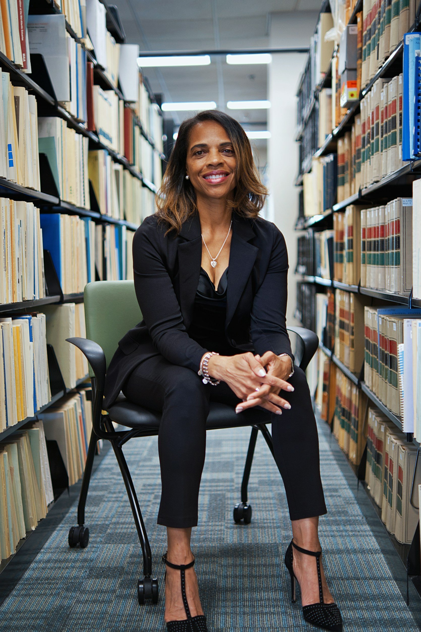 black woman executive sitting on a chair with her hands folded in front of her with medical files on a bookcase in the background
