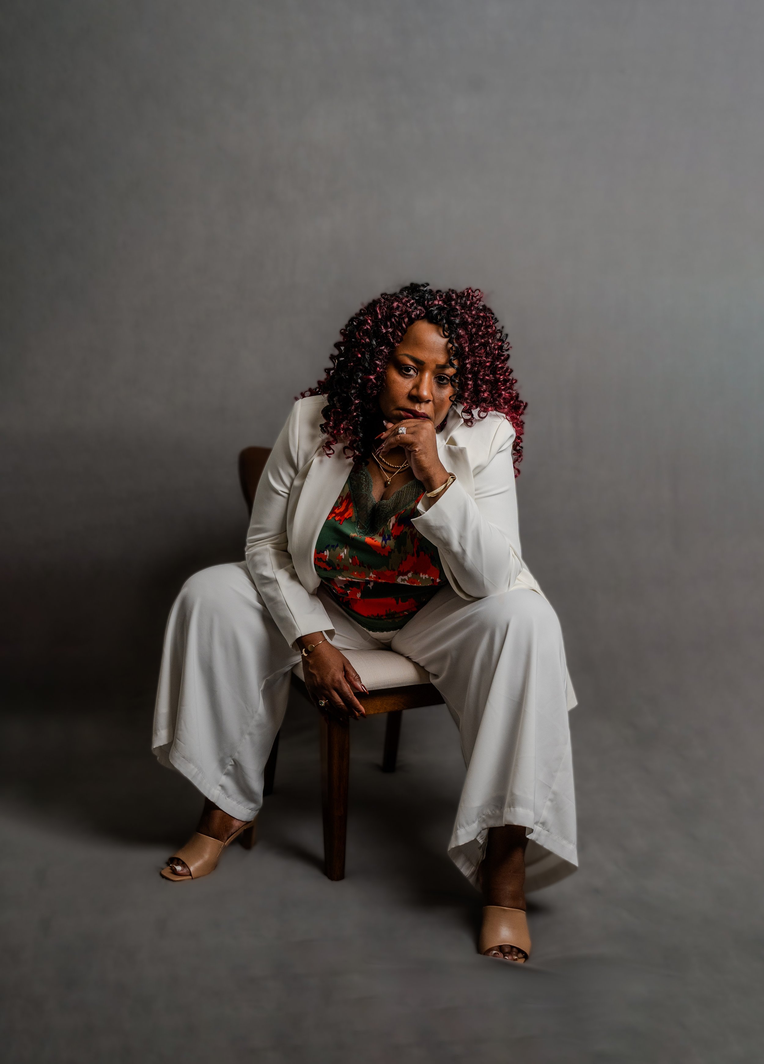 A woman with curly, dark red hair styled in ringlets, wearing a white pantsuit and high heels, sitting on a chair against a plain gray background, with a serious expression and her hand resting near her chin.