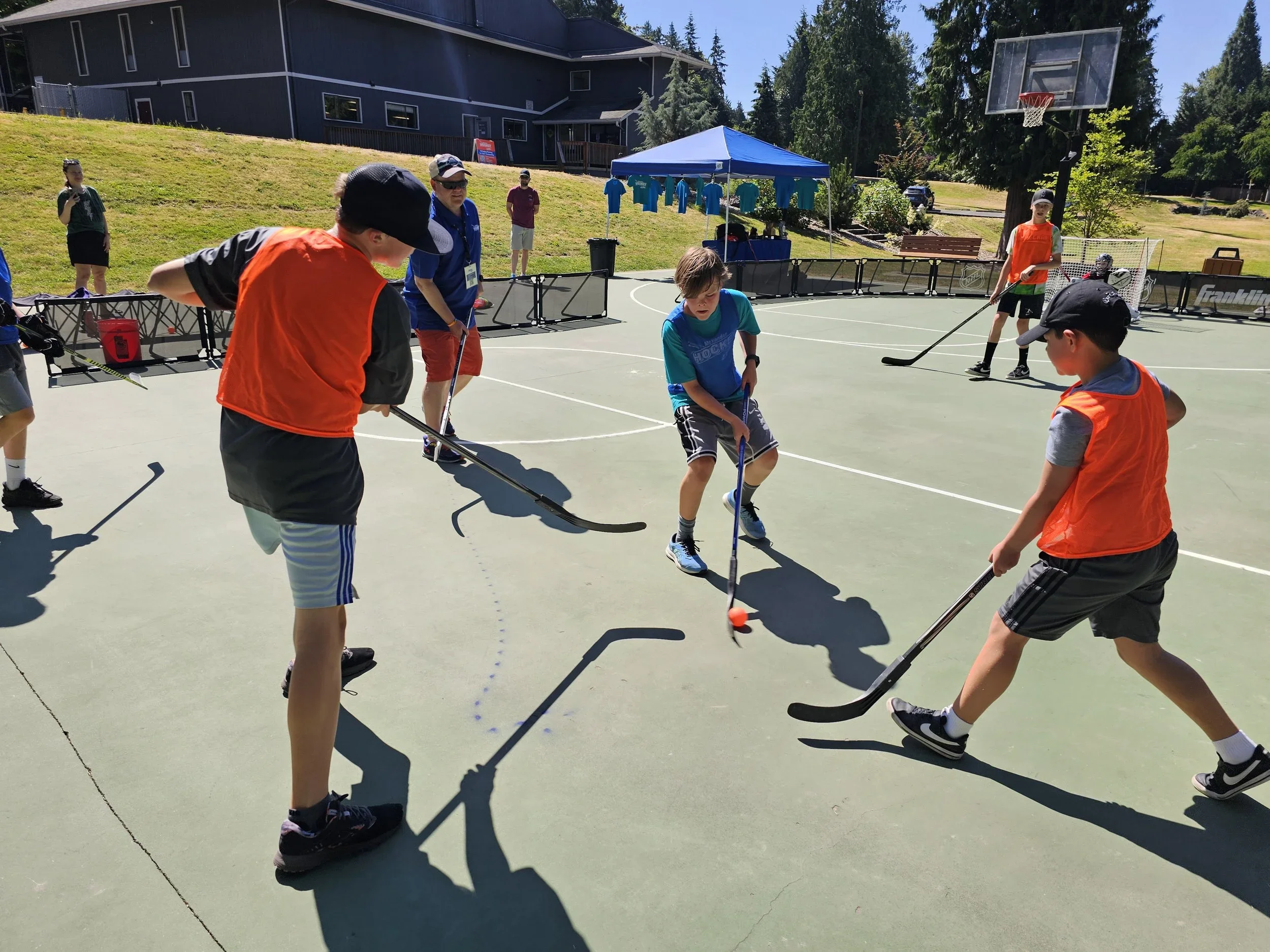 Young boys playing street hockey on an outdoor court with a blue tent and a basketball hoop in the background during a sunny day.