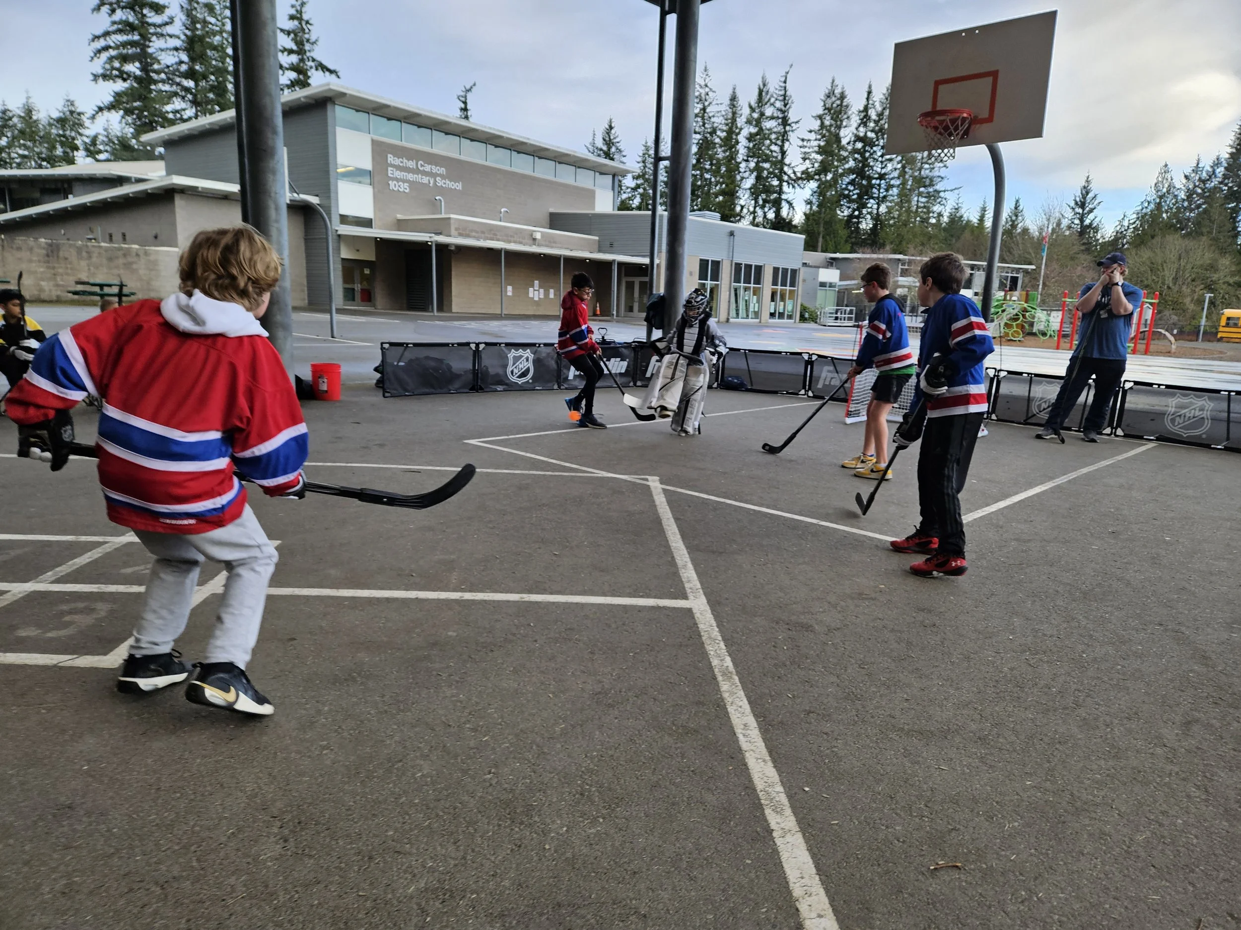 Group of children in hockey jerseys playing street hockey on a school playground.