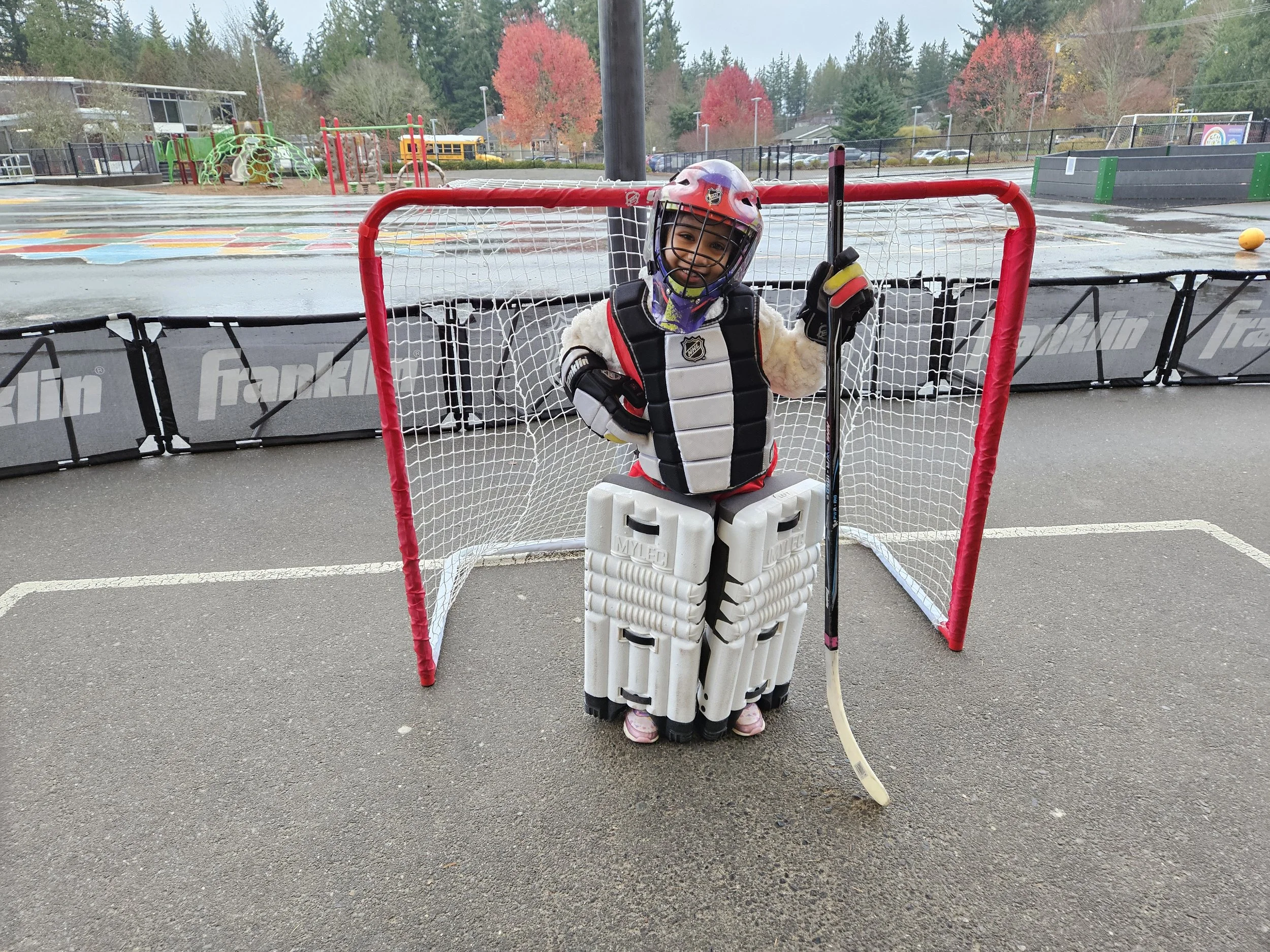 Young hockey goalie in full gear standing in front of a small hockey goal on in an outdoor area, with a playground and colorful trees in the background.