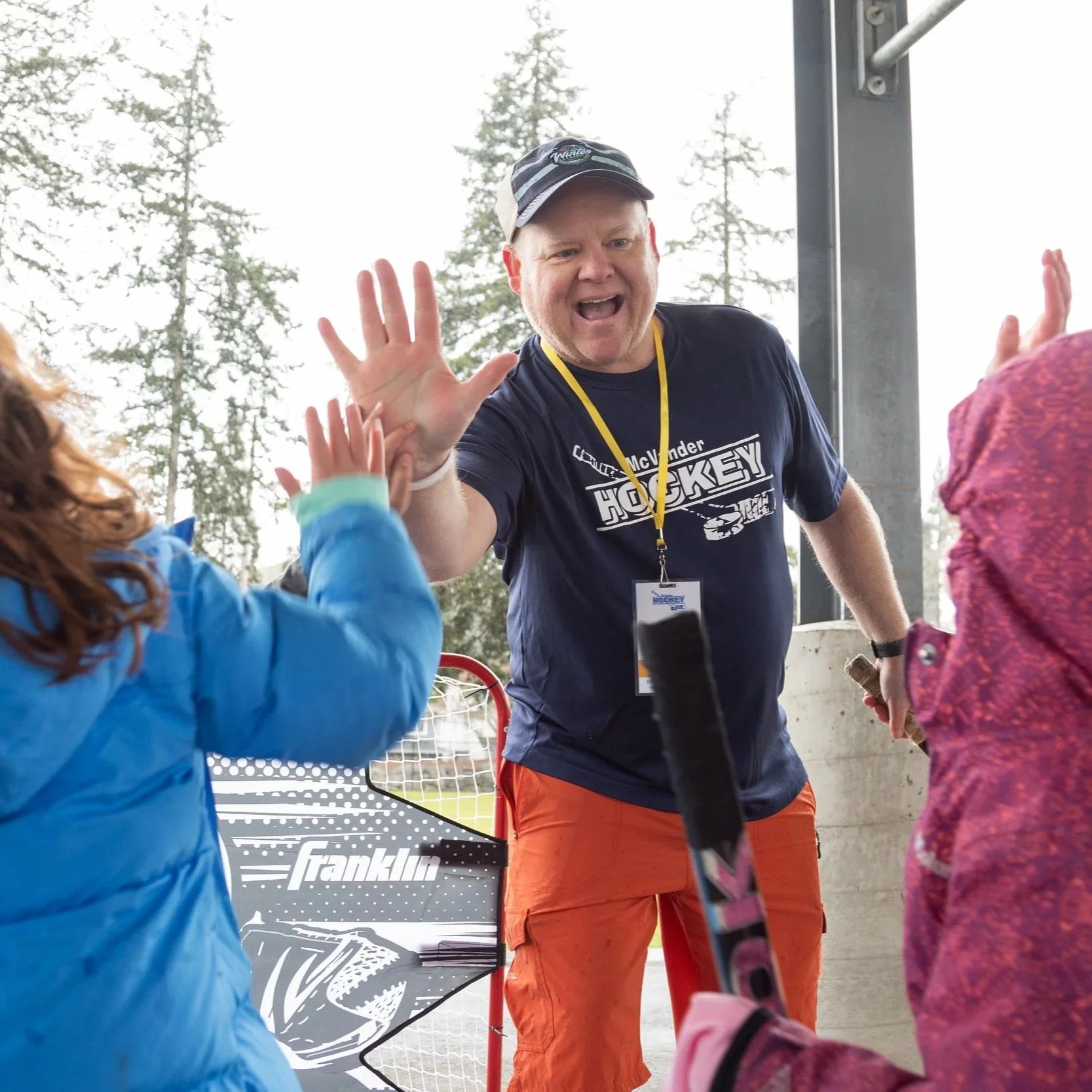 A man wearing a navy blue Hockey shirt, orange pants, and a cap high-fiving two children in outdoor sports attire near a hockey net.