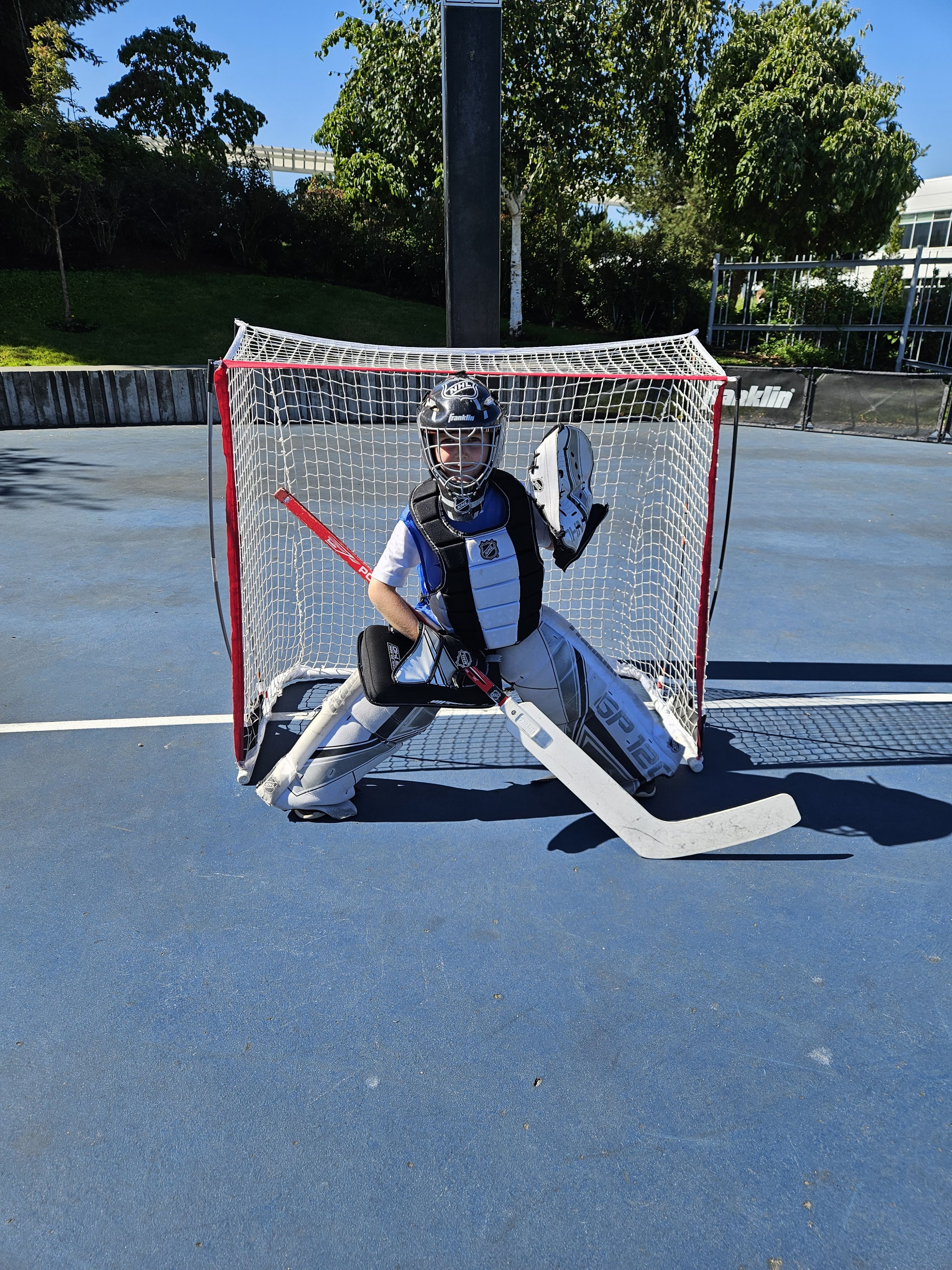A young hockey goalie in full gear, including a helmet, chest protector, and leg pads, is crouching in front of a hockey net on an outdoor blue rink, holding a goalie stick and wearing a catch glove, prepared for a shot.