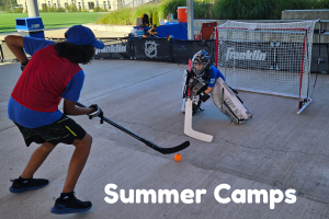 Two boys playing street hockey near a goal on a skate park with concrete surface, surrounded by a fence and summer camp signs.