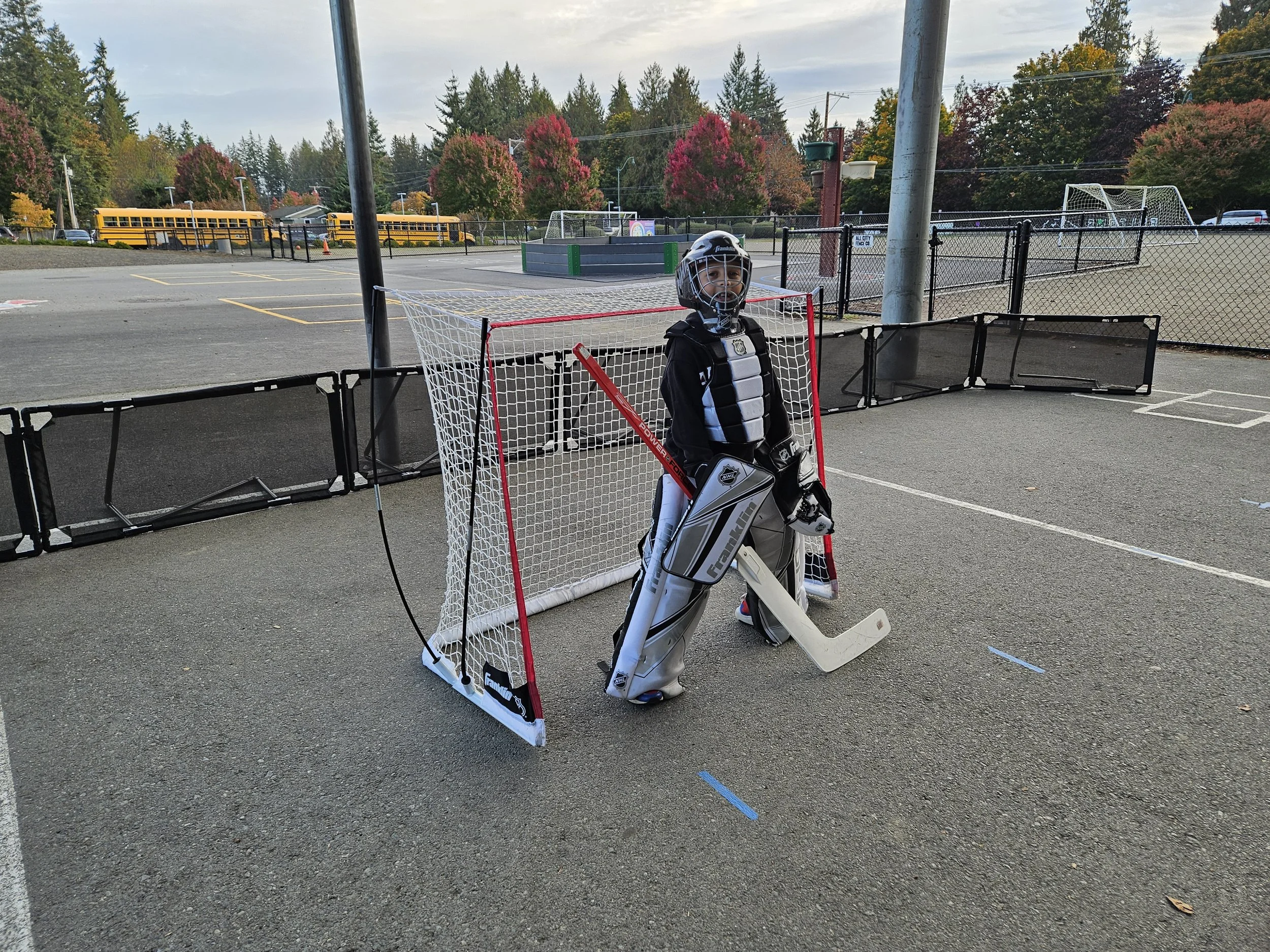 A young hockey goalie in full gear standing inside a small hockey net on an outdoor parking lot. The background features trees with fall foliage, parked school buses, and a sports field.
