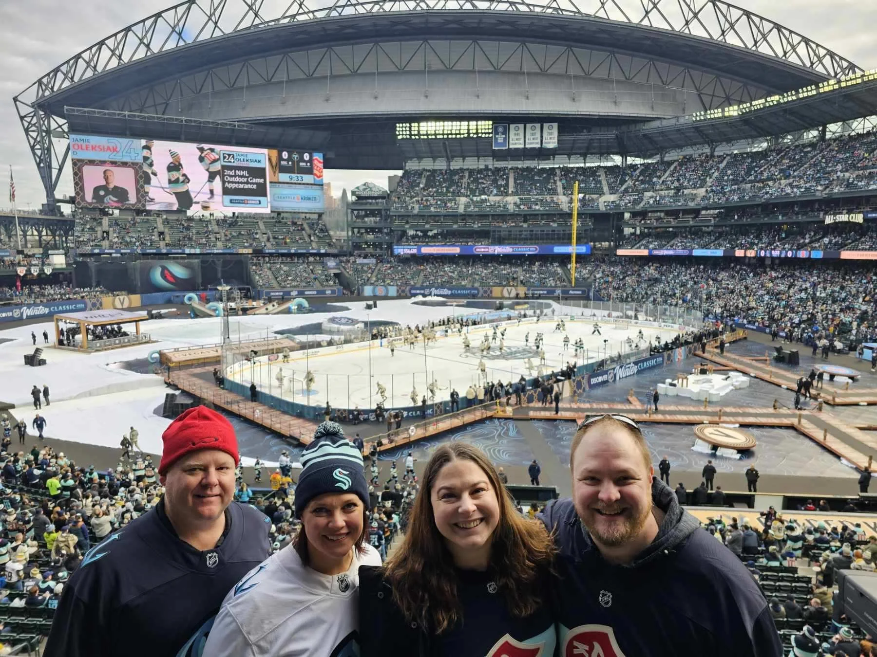 Four people smiling at a winter hockey game in a large stadium with many spectators. The ice rink is visible in the background with players warming up or practicing.
