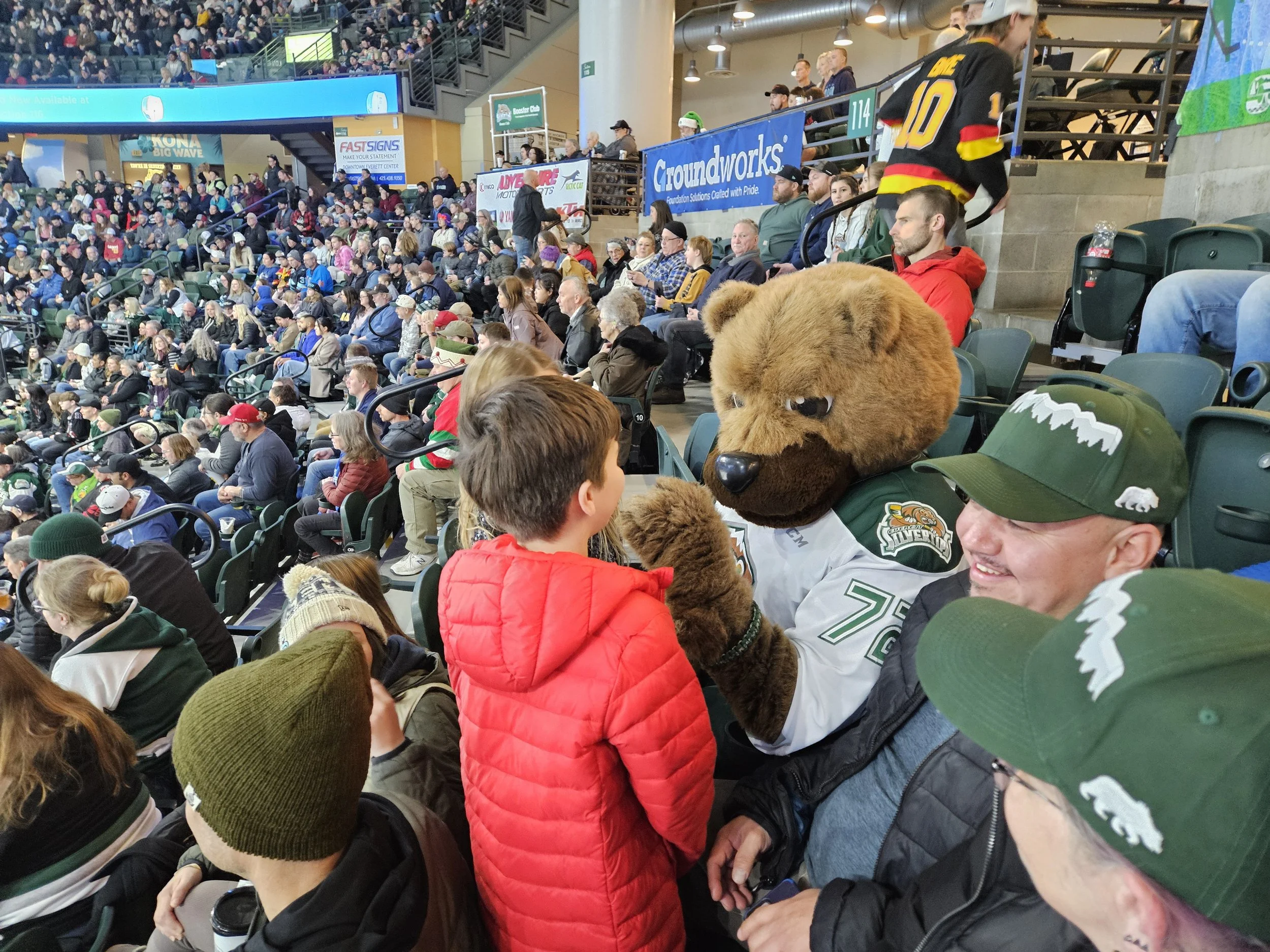 A large crowd of spectators at a hockey game, with a person in a bear mascot costume talking to a young boy in a red jacket in the foreground. The mascot is wearing a team jersey and appears to be shaking hands or high-fiving the boy.
