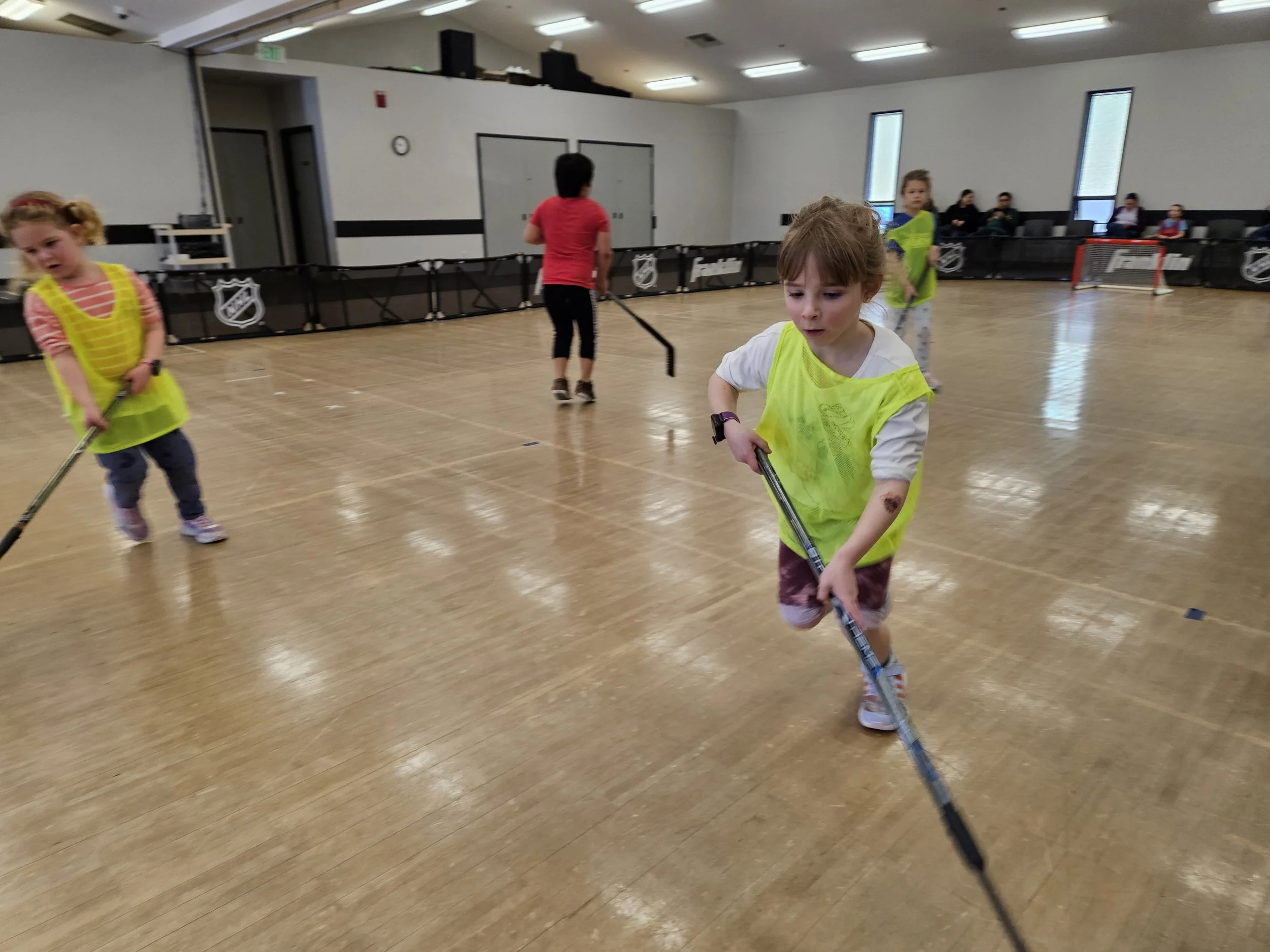 Children playing hockey in an indoor rink, wearing bright yellow vests, with spectators seated along the sides.