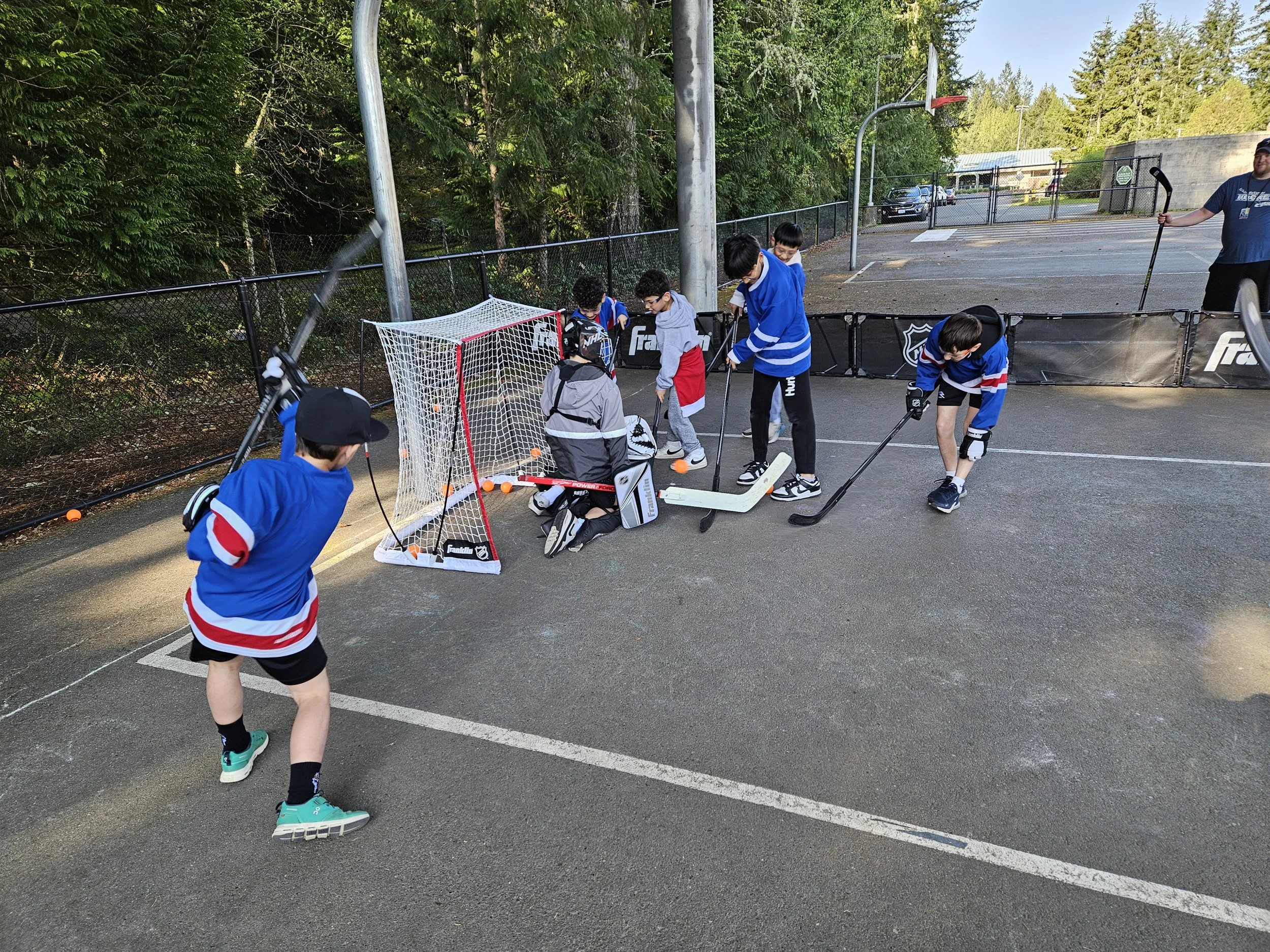 Children playing street hockey on an outdoor asphalt court with hockey sticks, puck, and small goal net, surrounded by a fence and trees.