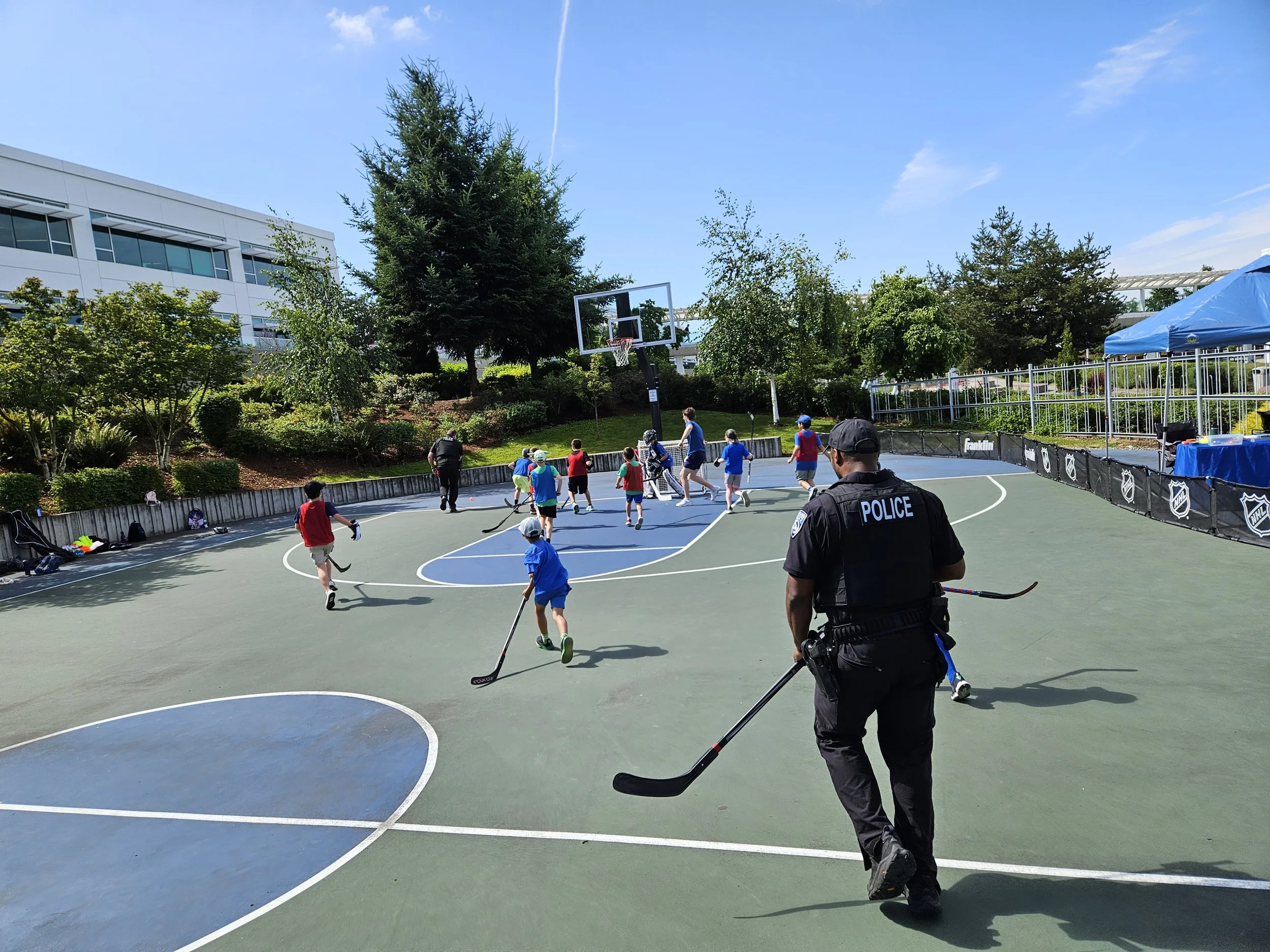Police officer watches children playing hockey on an outdoor basketball court on a sunny day.