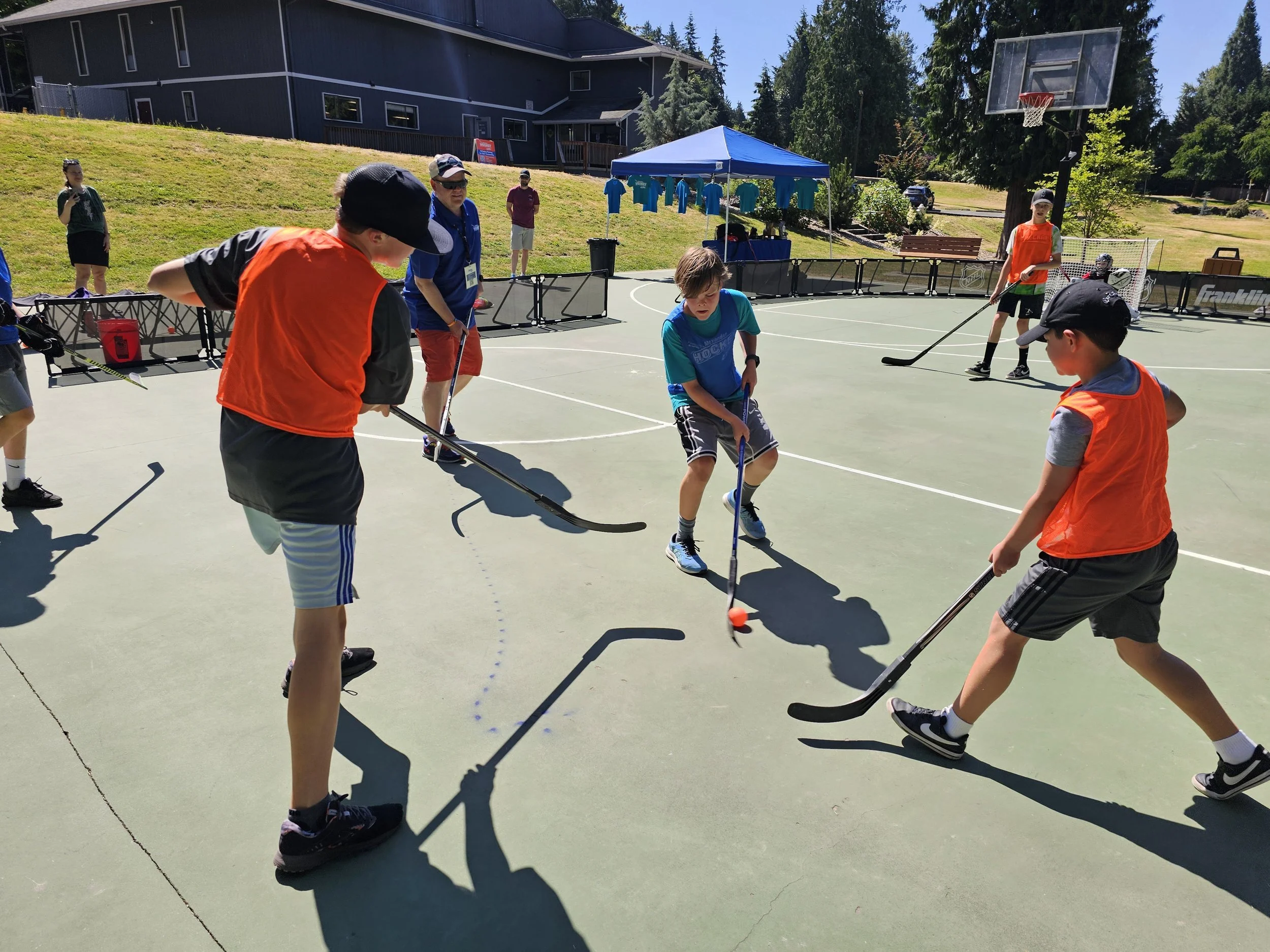 Children playing street hockey on an outdoor court under a sunny sky, with a blue canopy and a basketball hoop in the background.