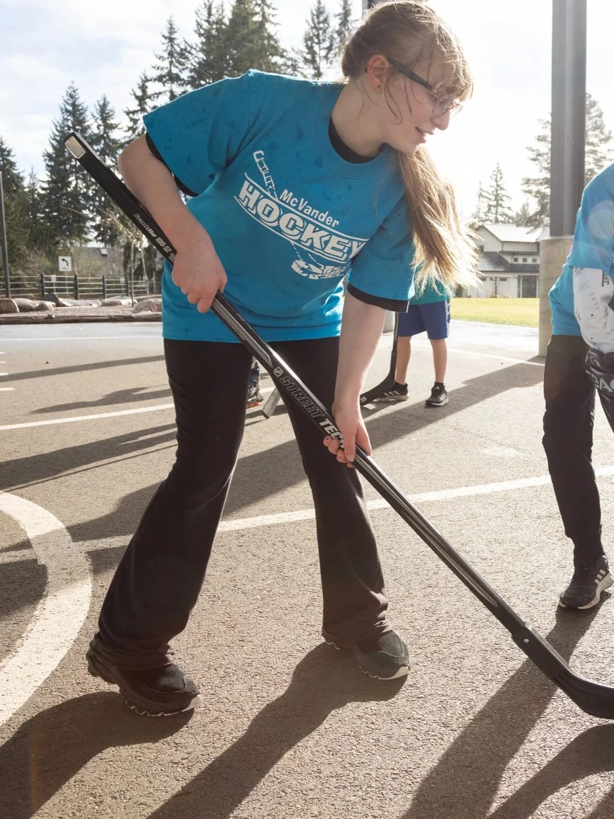 A girl in a blue hockey jersey and black pants holding a hockey stick on an outdoor pavement.