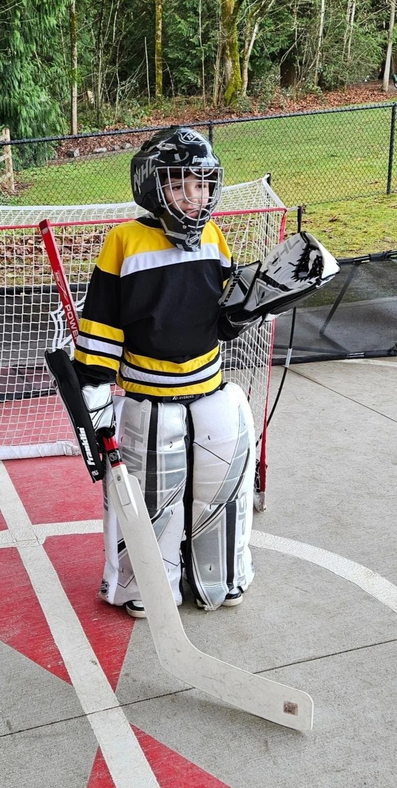 Young boy dressed as a hockey goalie standing on an outdoor hockey rink, wearing a black and yellow hockey jersey, goalie pads, helmet, and holding a hockey stick, with a hockey net in the background.