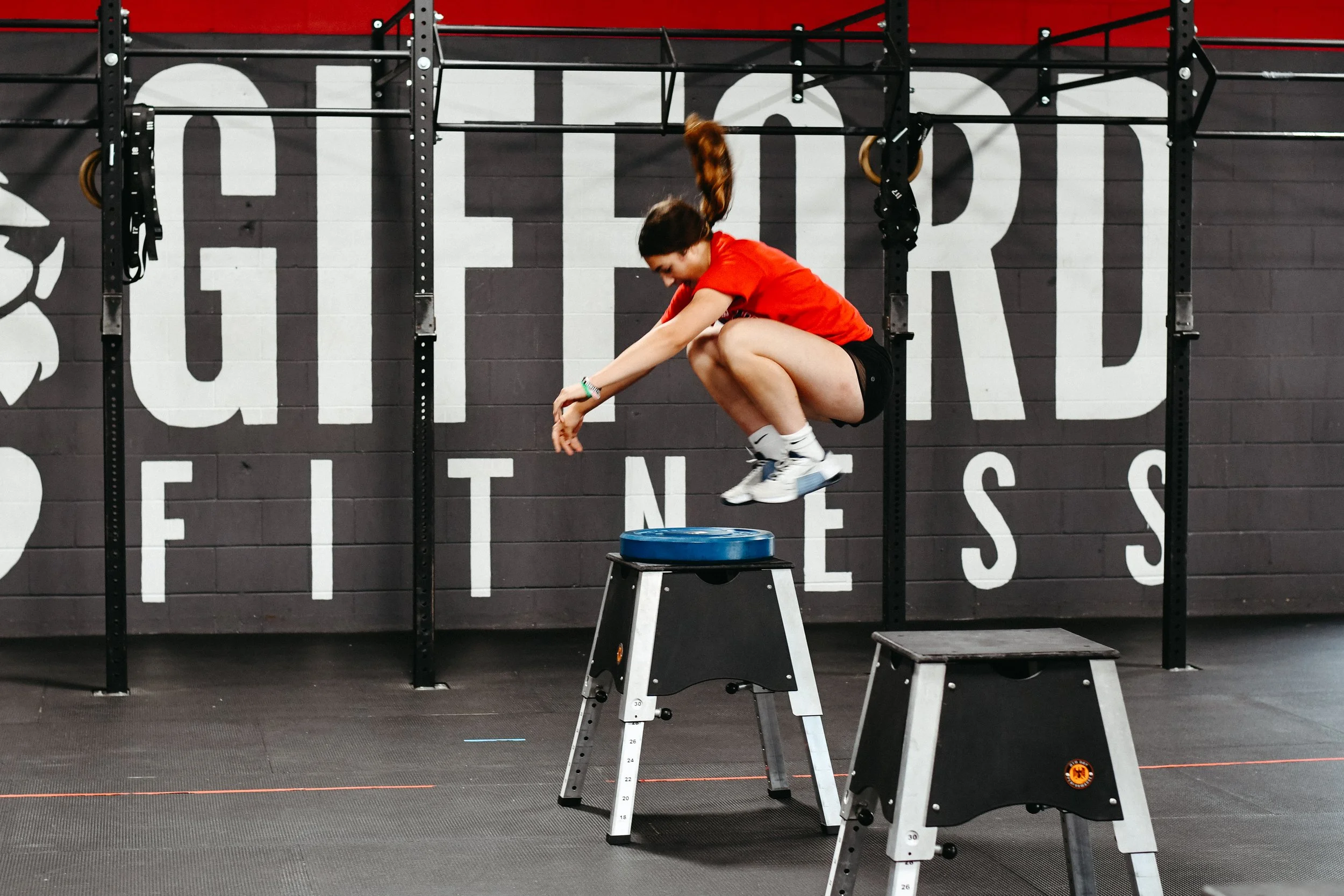 A woman jumping onto a plyometric box in a gym, with a mural wall behind her that says 'GIFFORD FITNESS'